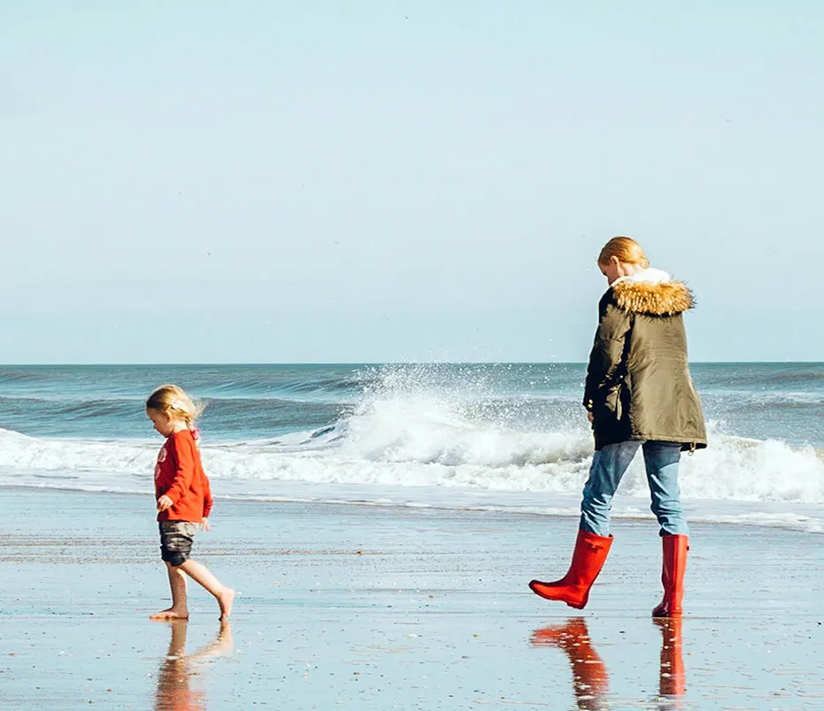 An adult and a child walk along a beach near the ocean. The adult wears a green coat, blue jeans, and red boots. The child, who has long blond hair, wears a red shirt and shorts and is barefoot.