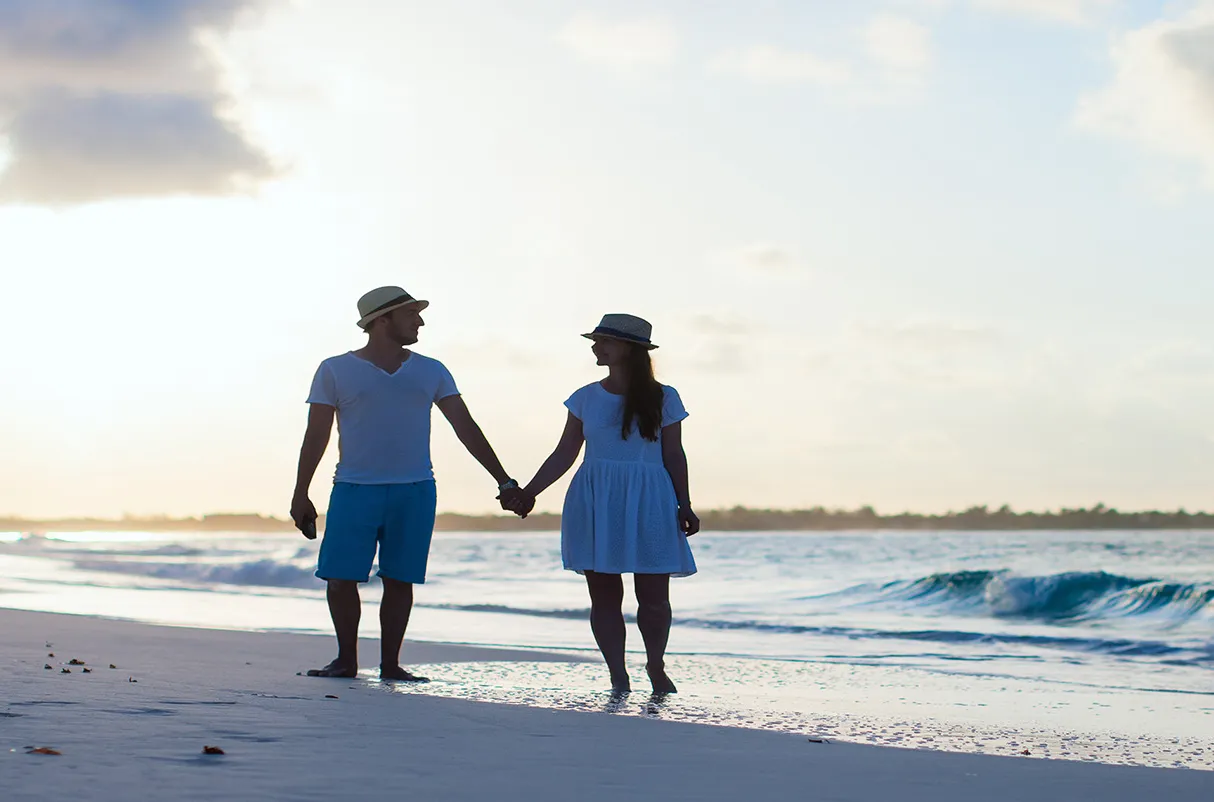 A couple, both wearing hats, holding hands and walking along the shore of a beach during sunset.