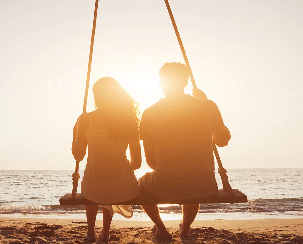Two people sit side by side on a swing by the beach at sunset, facing the ocean with the sunlight creating a soft glow around them.