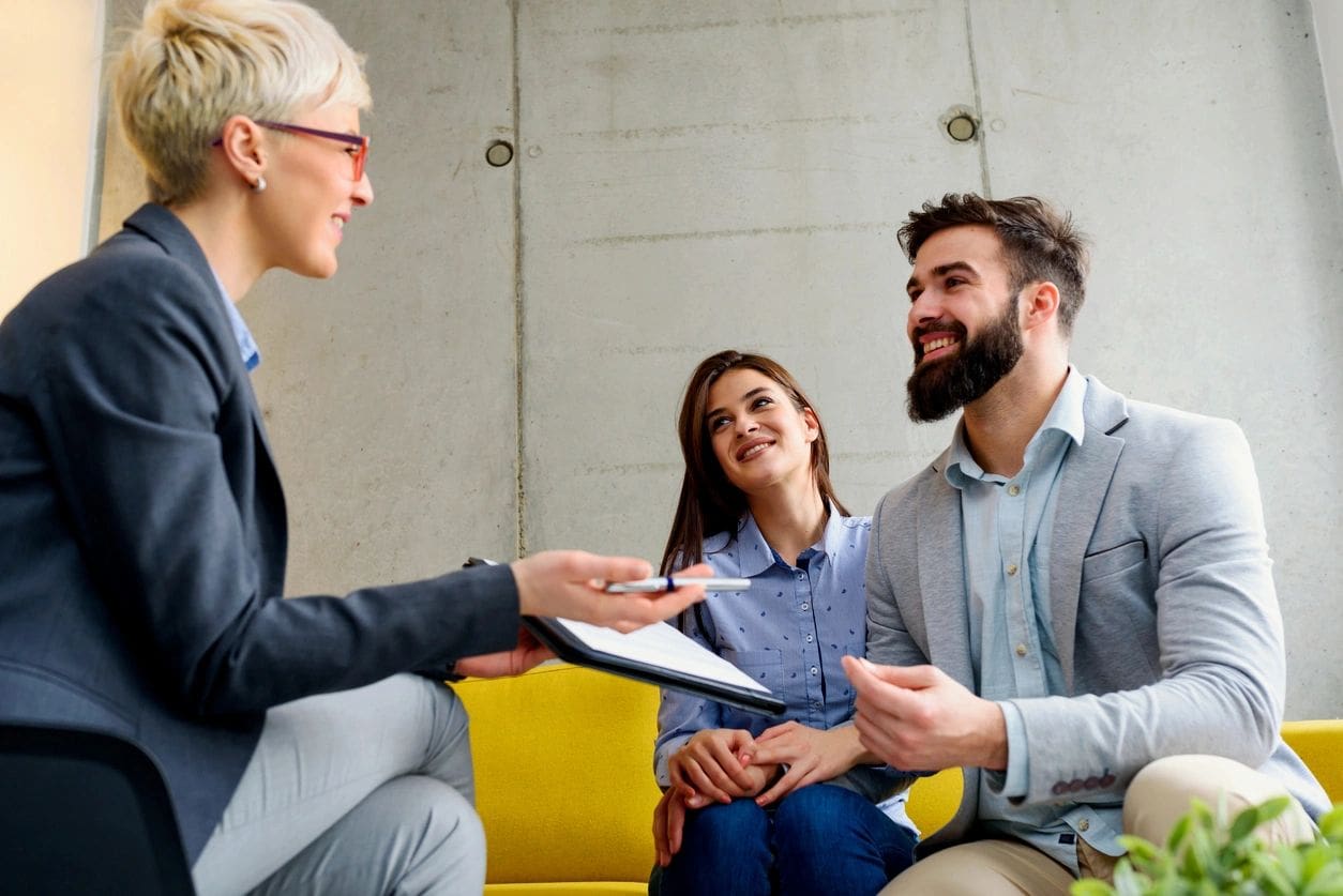 A businesswoman with a clipboard discusses with a smiling couple seated on a yellow couch in a modern office setting.