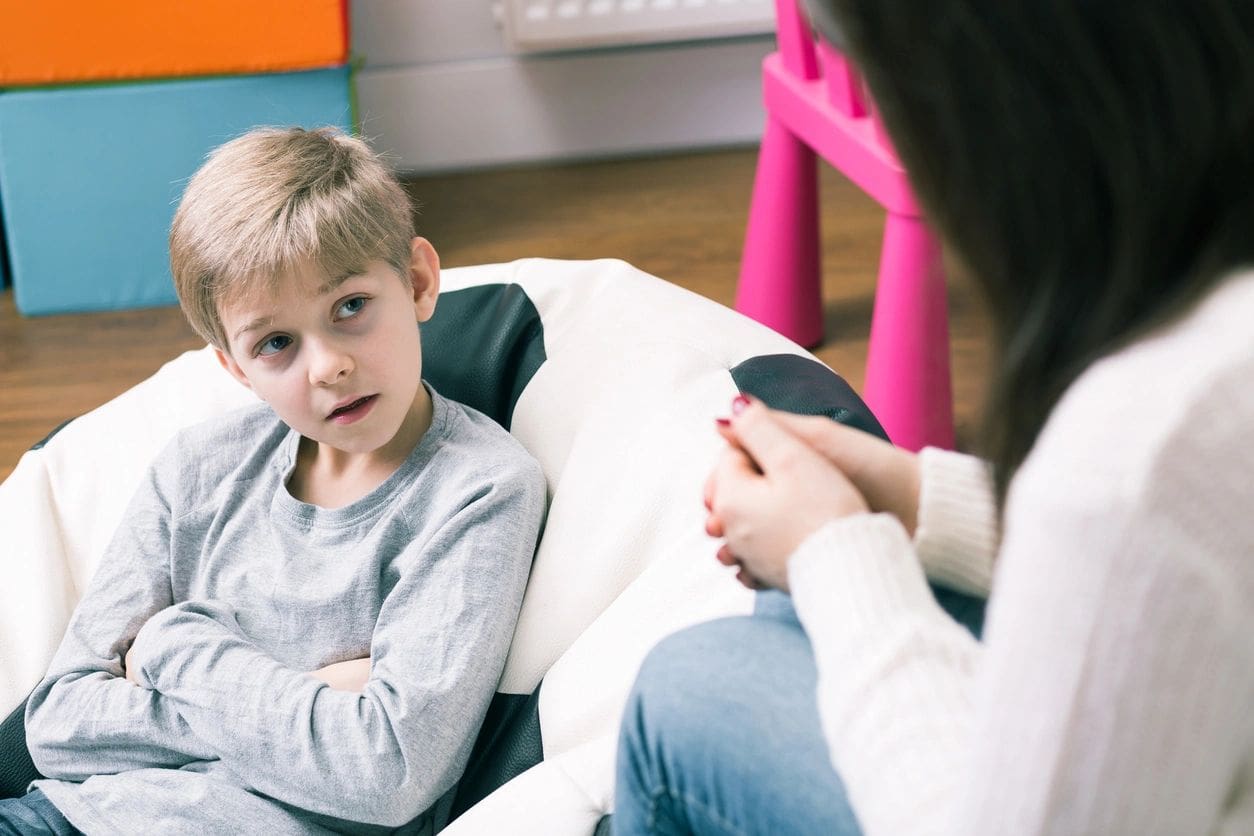 A child sits on a bean bag chair with arms crossed, listening attentively to a person in a white sweater sitting across from them.